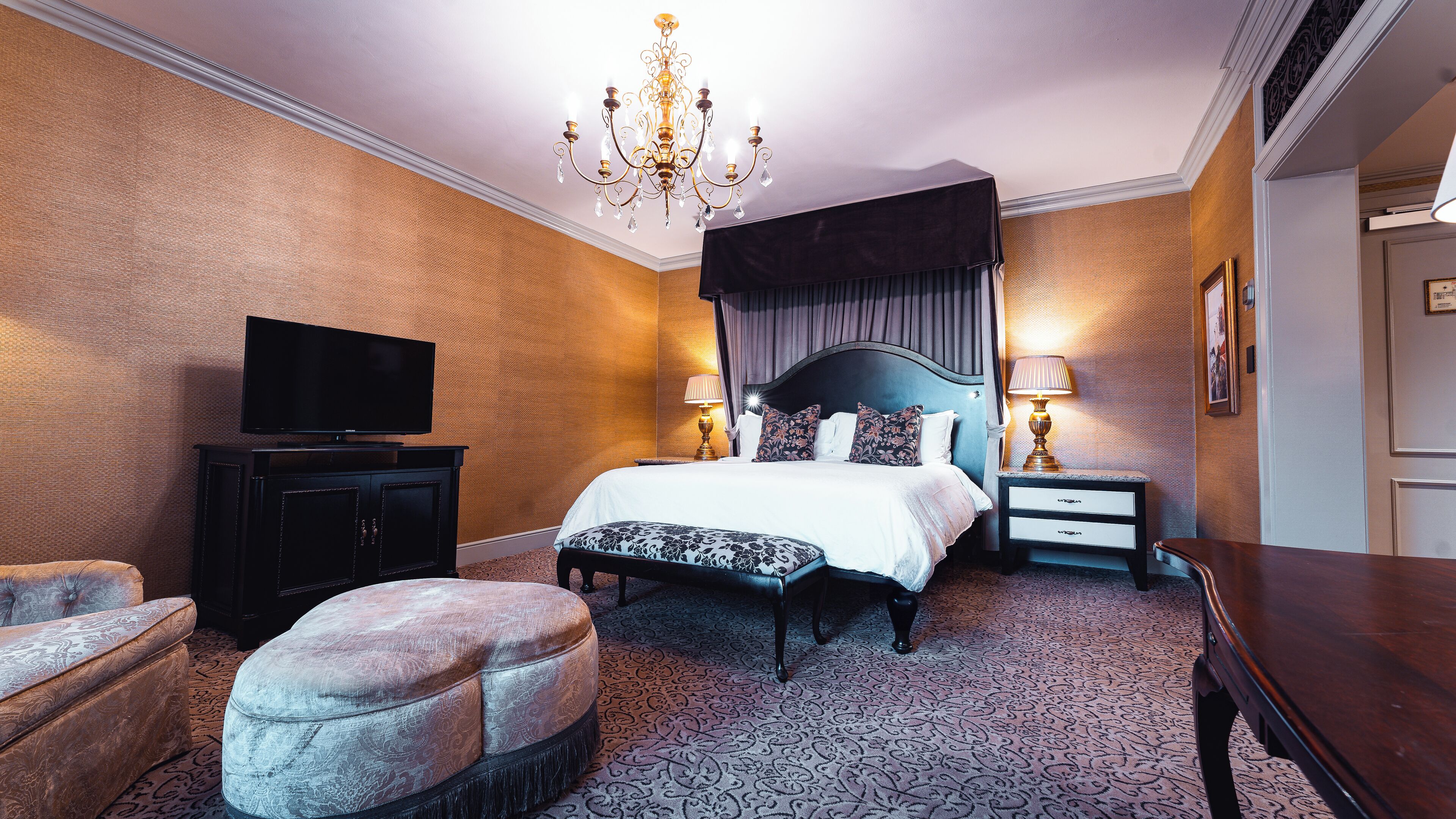 A hotel room with a large bed, a television, a chandelier, a nightstand, and a patterned carpet.