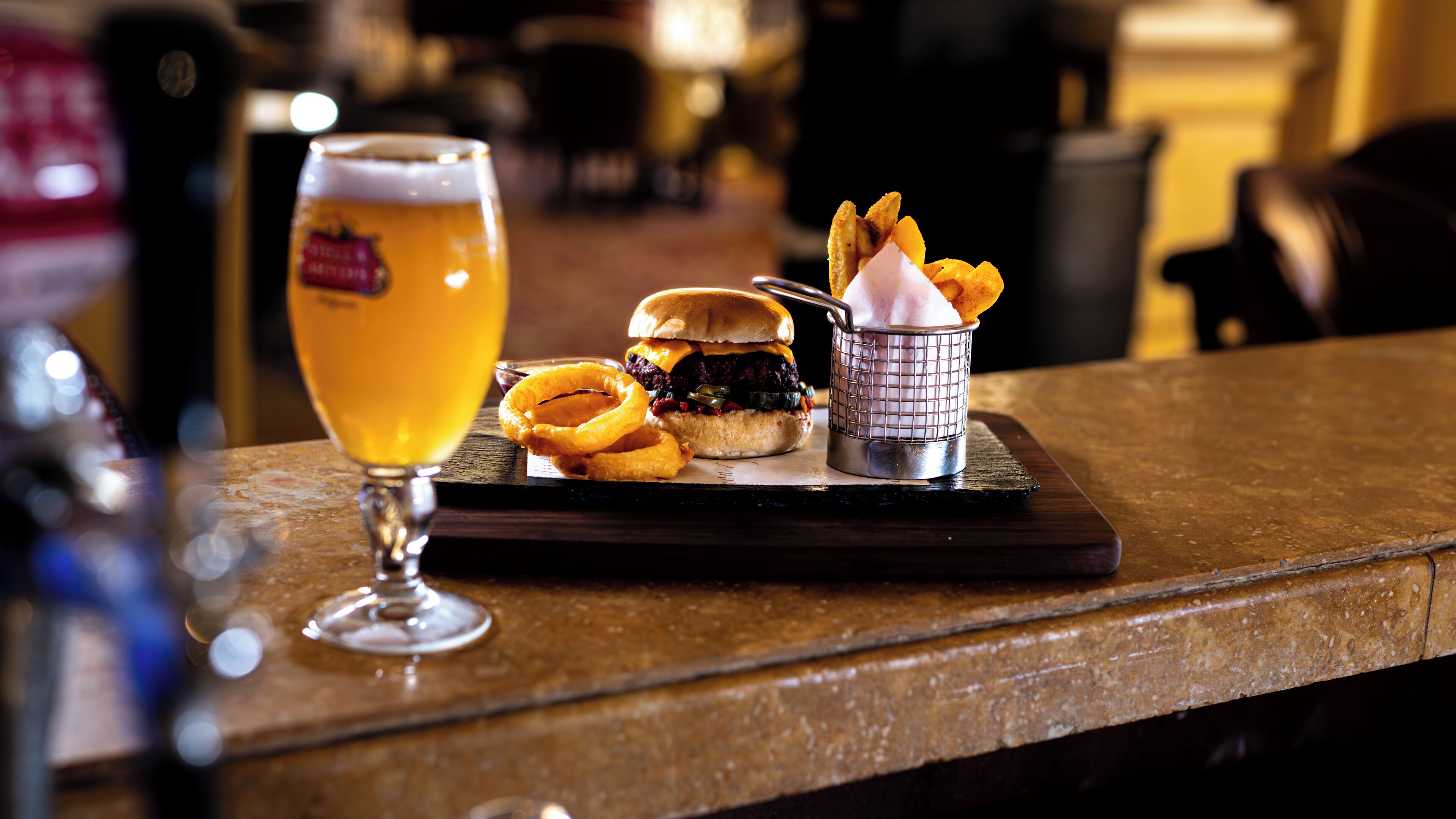 En barscene med et glas øl, en burger, løgringe og en portion pommes frites på en træplanke.