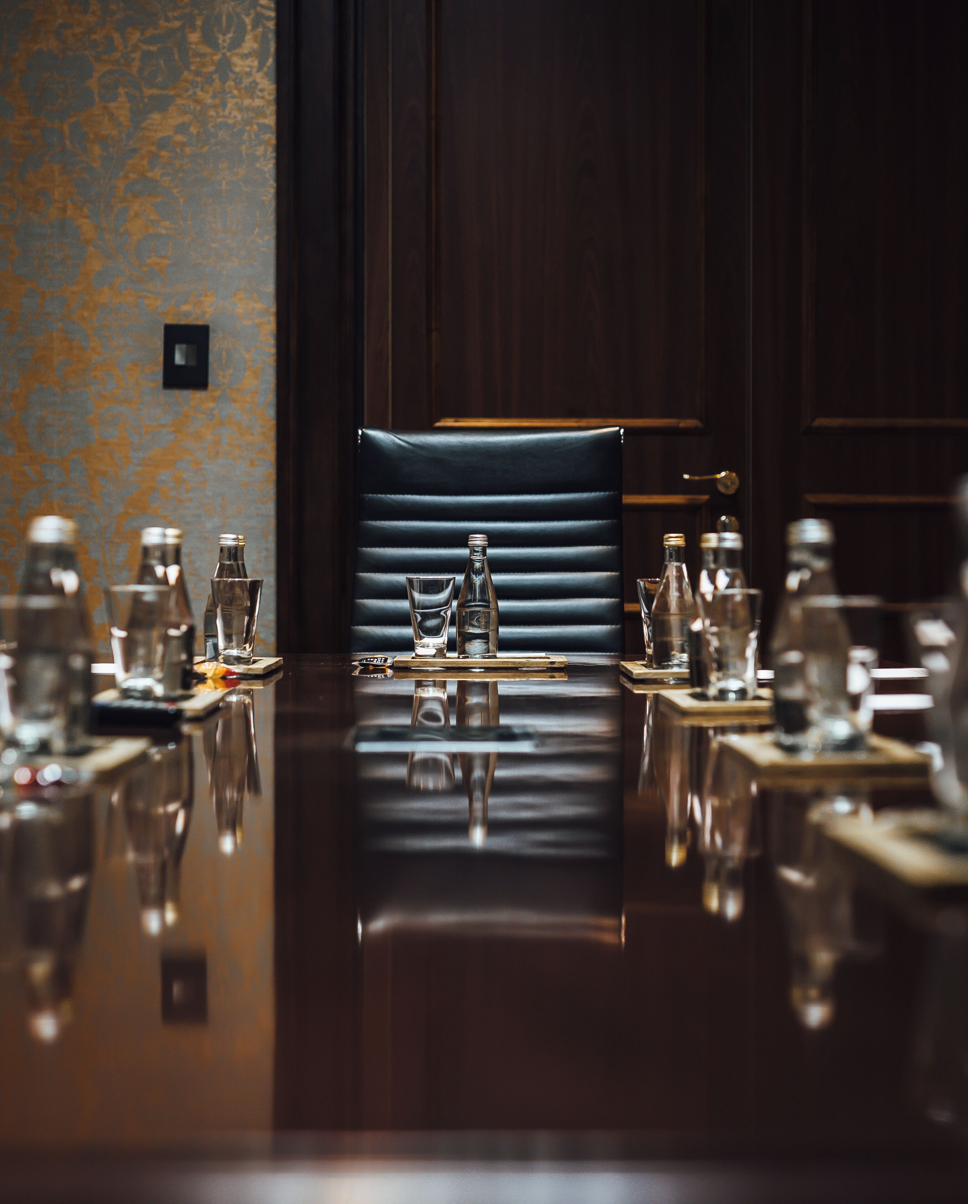 A conference table with chairs, water bottles, and glasses, set in a room with patterned wallpaper and wooden paneling.