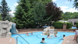 A swimming pool with people enjoying the water, surrounded by trees and a stone waterfall feature.