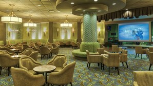 Um saguão de hotel com mesas redondas, cadeiras acolchoadas, um pilar grande e uma tela exibindo um filme.