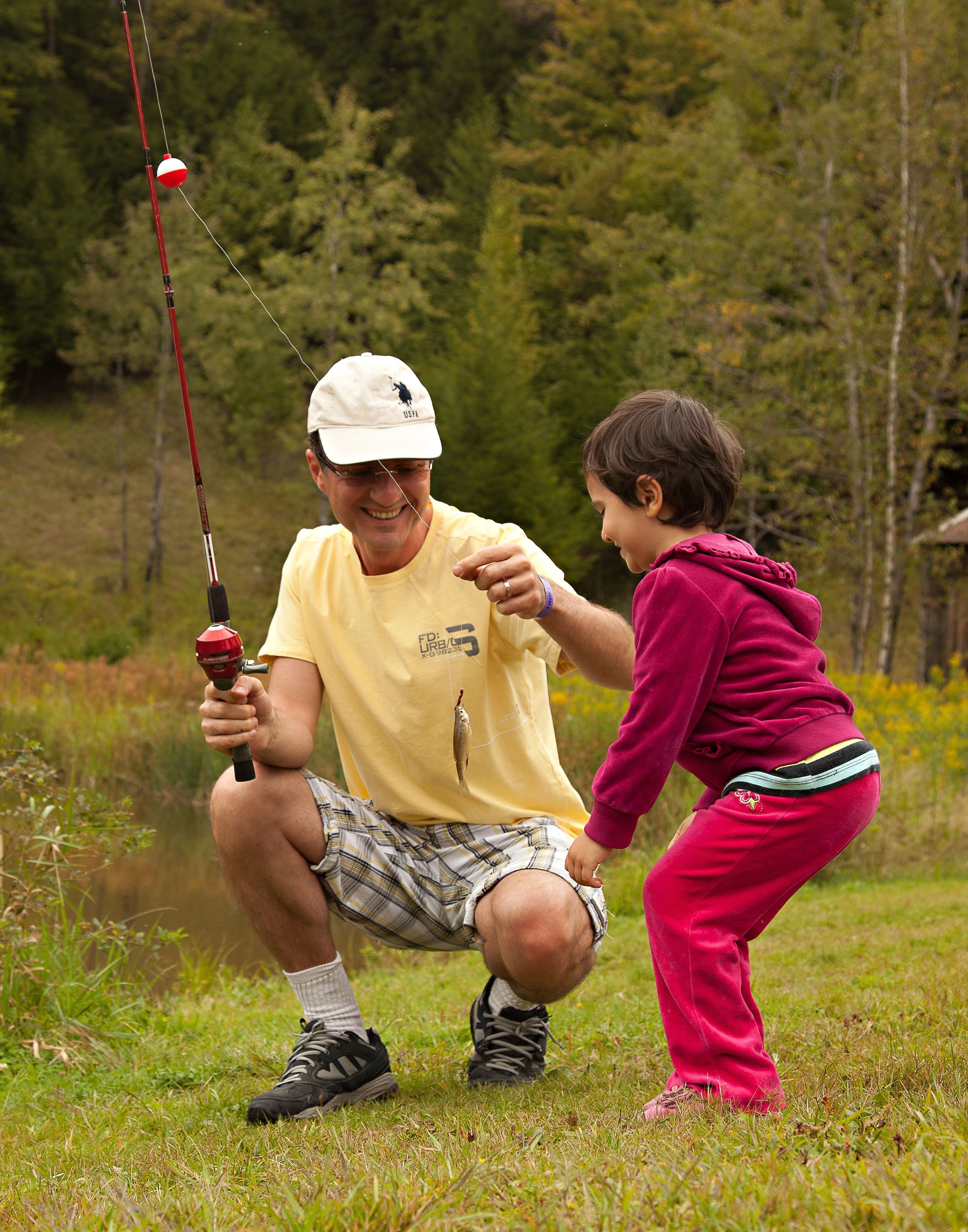 A man and a child fishing by a calm body of water surrounded by greenery.