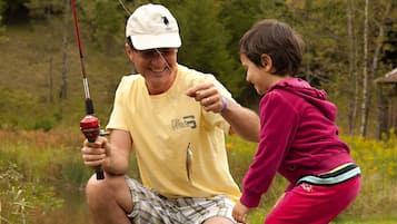 A man and a child fishing by a calm body of water surrounded by greenery.