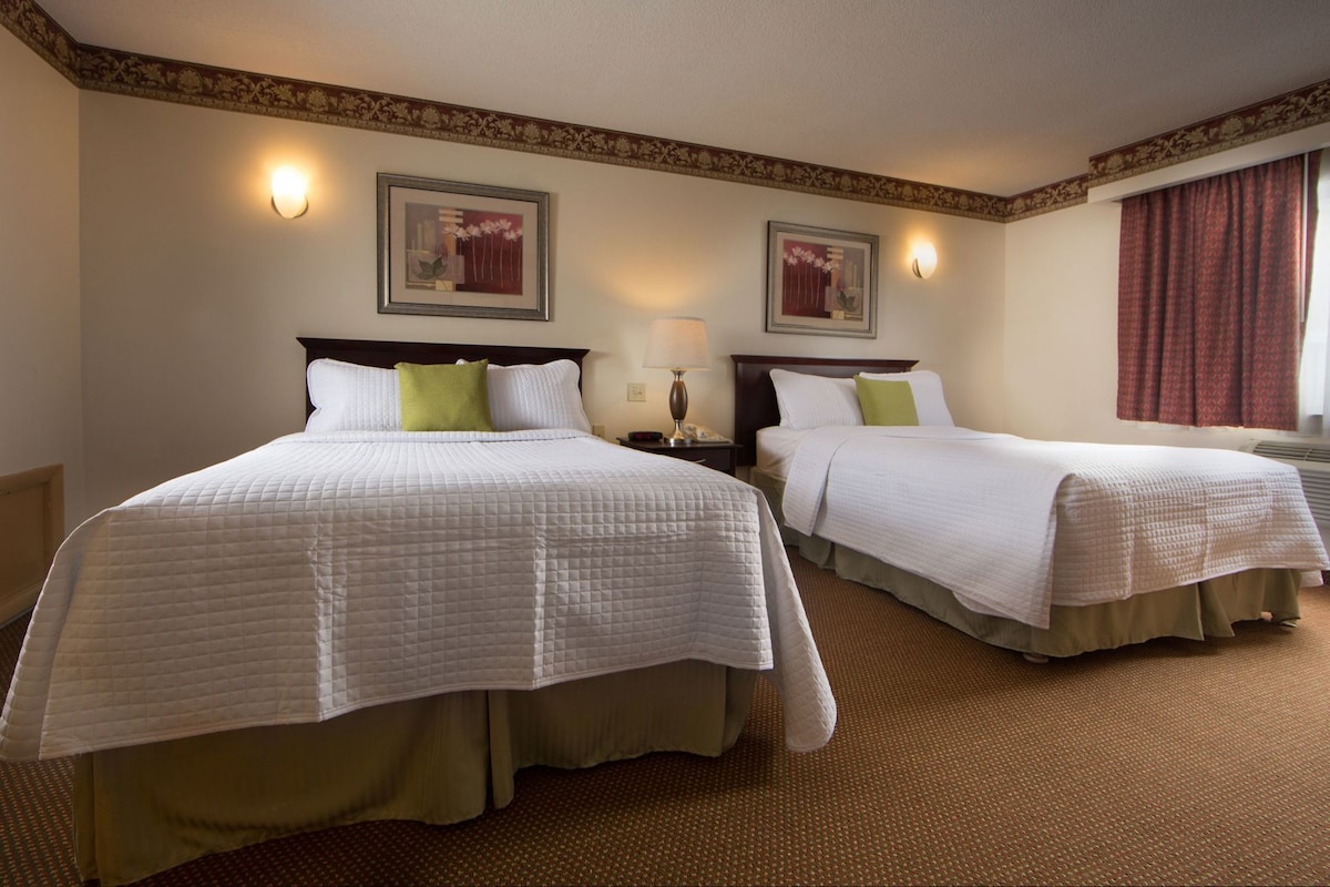 A hotel room with two beds, a nightstand with a lamp, a wall-mounted picture, and a window with curtains.