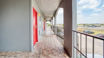 A covered balcony with a tiled floor, red doors, and a view of a parking lot and distant buildings.