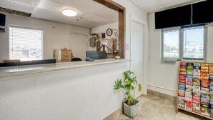 Zona de recepción con mostrador, planta en maceta, tablón de anuncios y ventana con persianas.