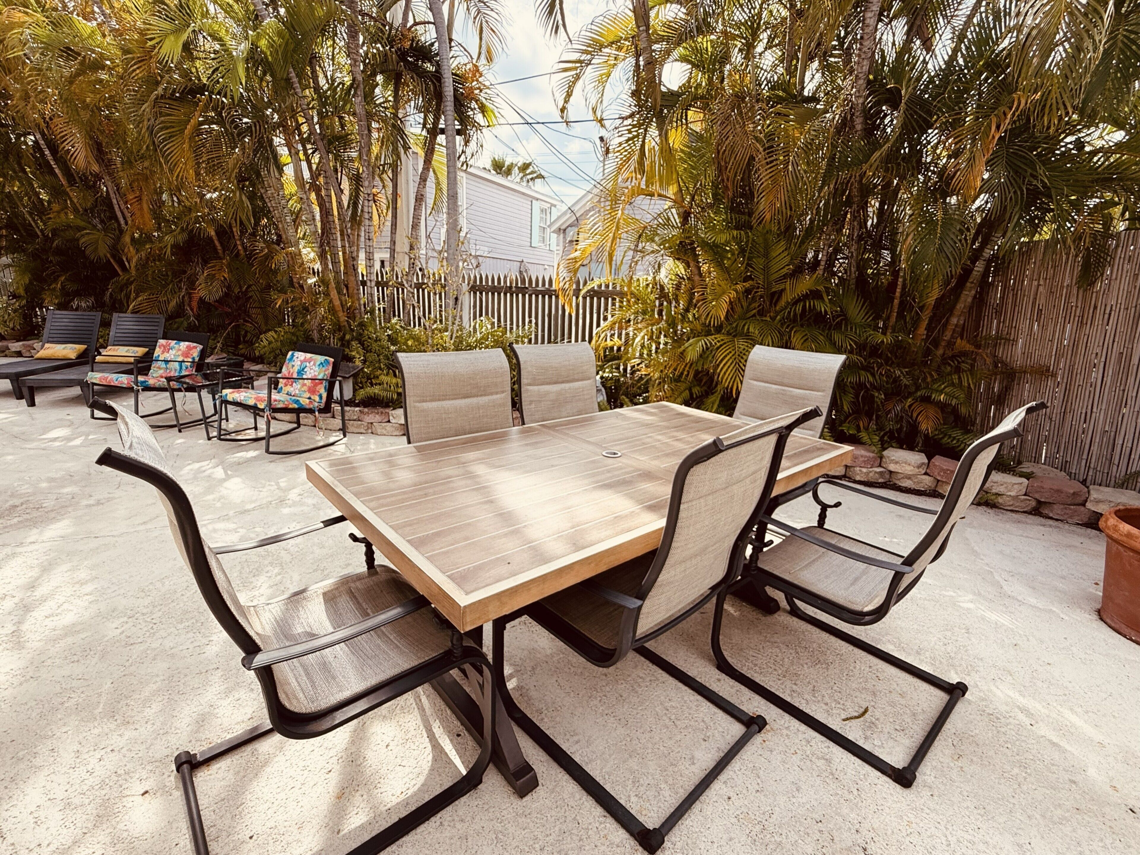 An outdoor patio with a wooden table and chairs, surrounded by lush greenery and a white picket fence.