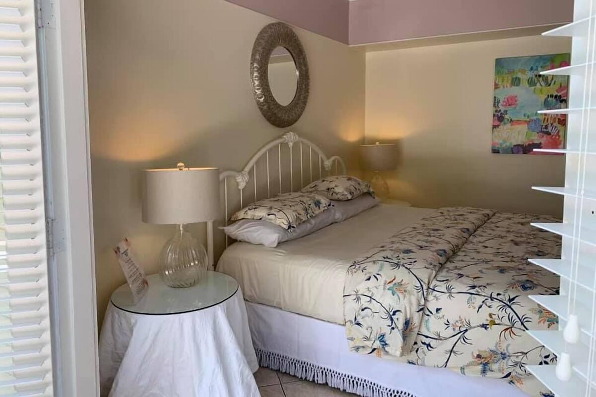 Ein Schlafzimmer mit einem Bett, einem Nachttisch mit Lampe, einem Wandspiegel und einem farbenfrohen Gemälde.