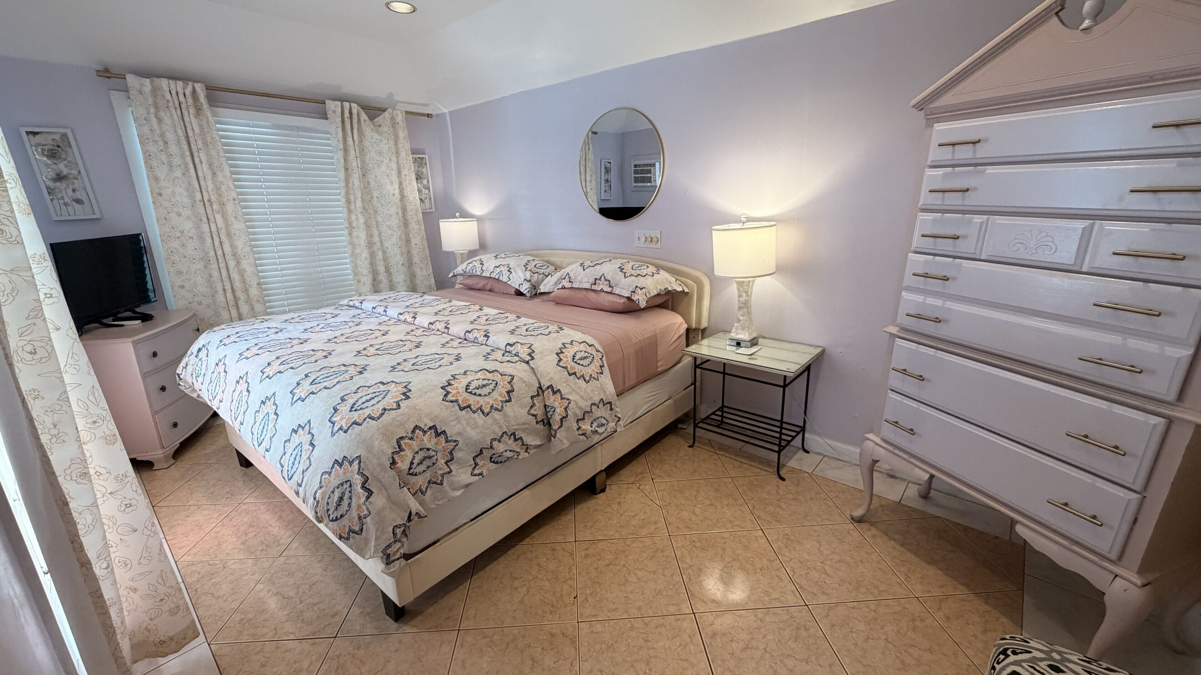 Un dormitorio con cama, mesitas de noche, cómoda, espejo y ventana con cortinas.