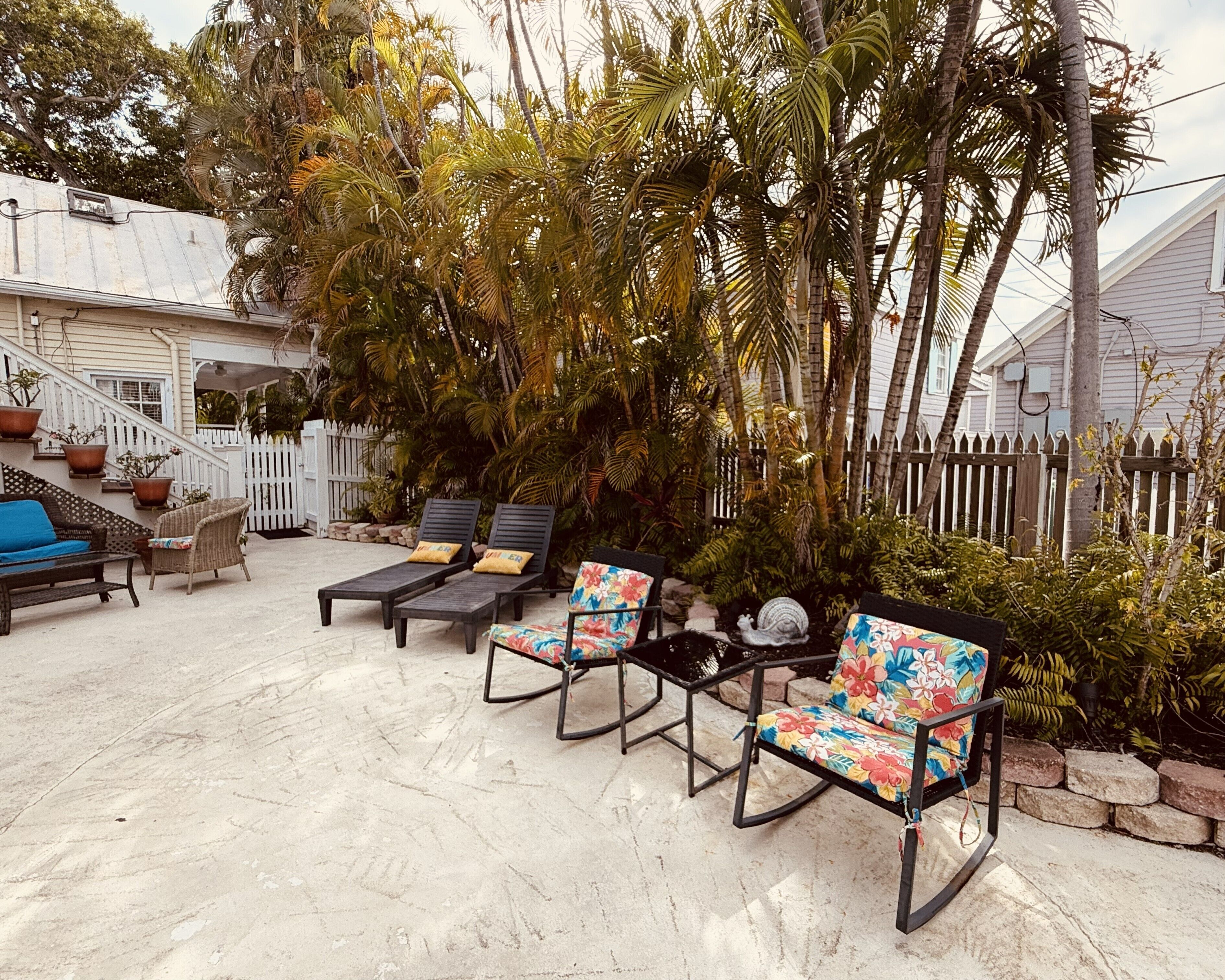 A patio with outdoor furniture, including lounge chairs and a sofa, surrounded by a white picket fence and lush greenery.
