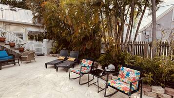A patio with outdoor furniture, including lounge chairs and a sofa, surrounded by a white picket fence and lush greenery.