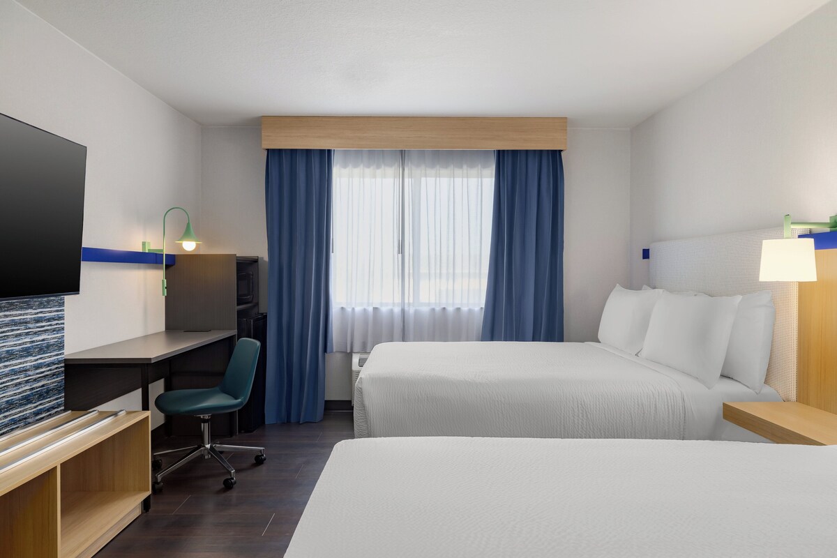 Ein Hotelzimmer mit zwei Betten, einem Schreibtisch, einem Stuhl, einem Fernseher und einem Fenster mit Vorhängen.