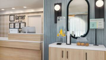 A hotel reception area with a wooden reception desk, framed artwork, a mirror, and a sign with amenities.