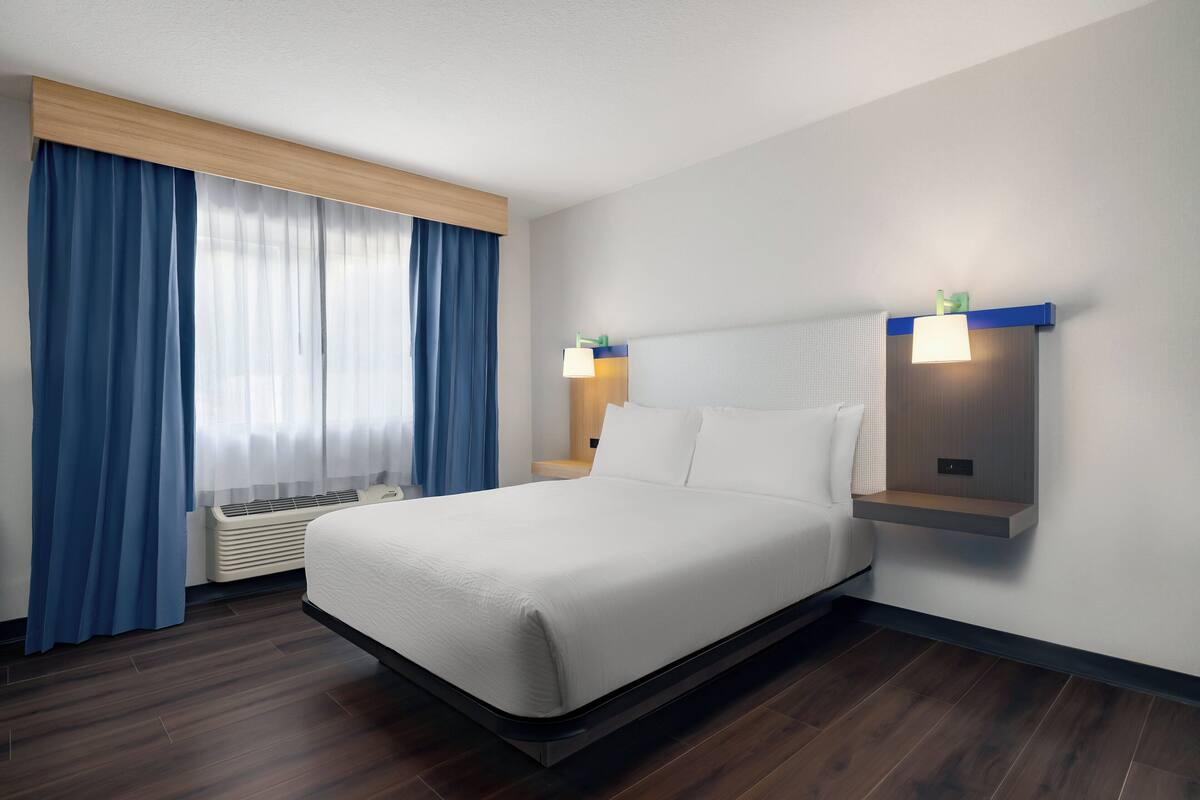 A modern hotel room with a large bed, blue curtains, a wall-mounted light fixture, and a built-in shelf.