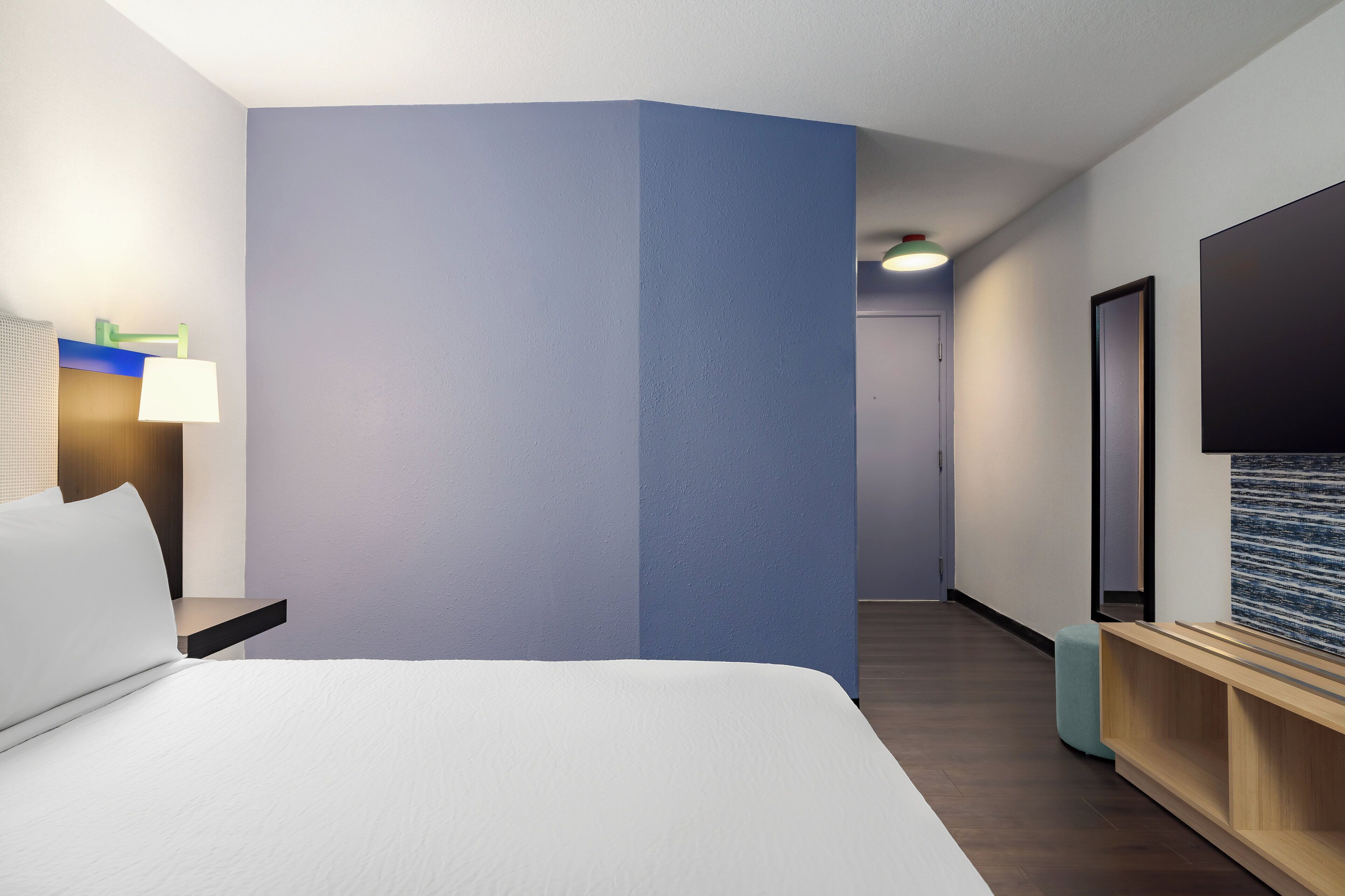 A hotel room with a bed, a nightstand, a lamp, a TV, and a wooden bench.