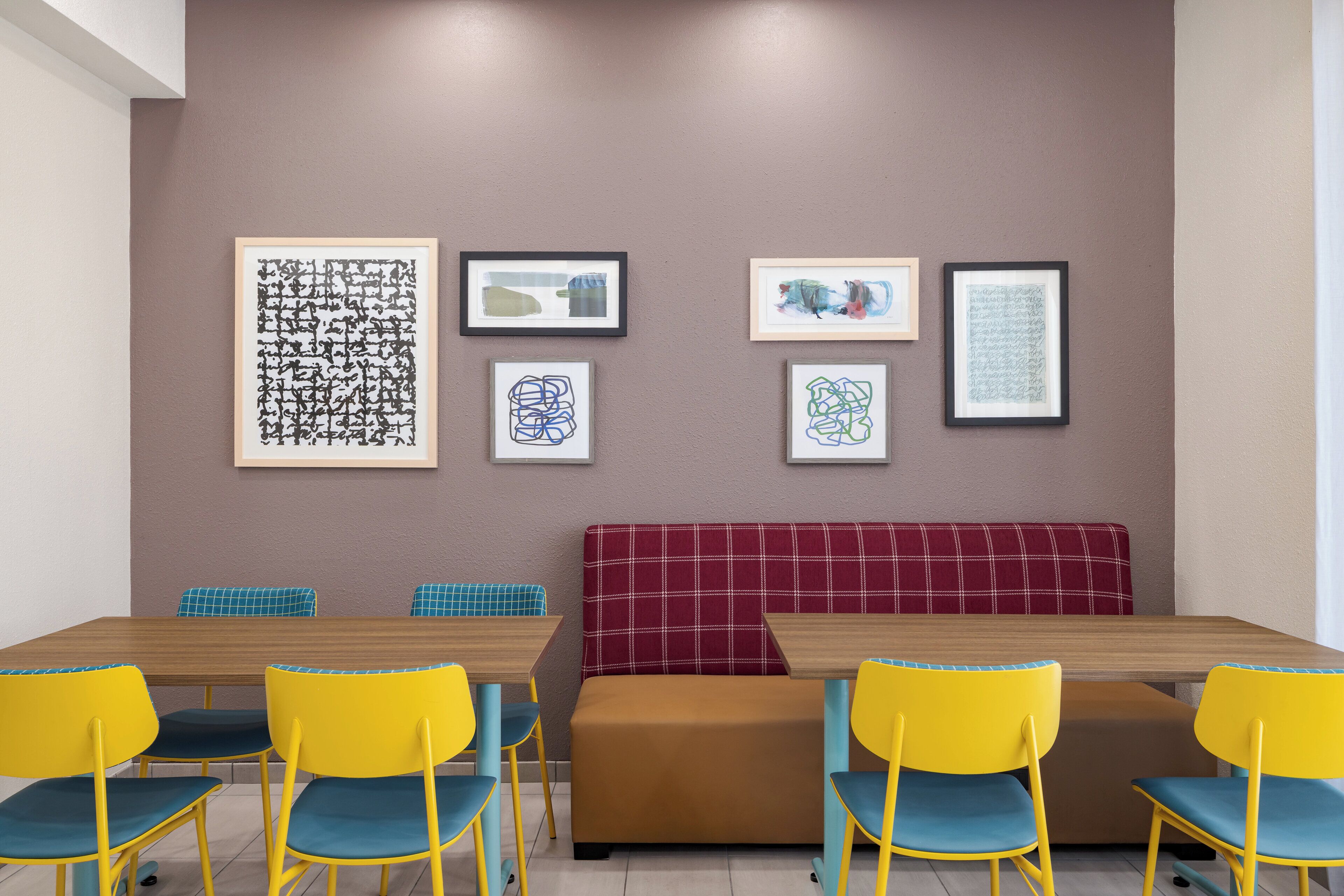 A dining area with a wooden table, yellow and blue chairs, a red and brown bench, and framed abstract art on the wall.