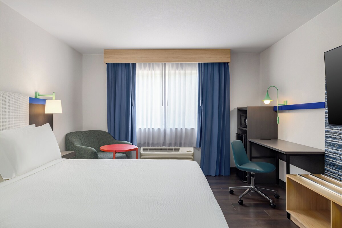 Ein Hotelzimmer mit einem Bett, einem Schreibtisch, einem Sessel, einem kleinen Tisch, einem Fernseher und einem Fenster mit Vorhängen.