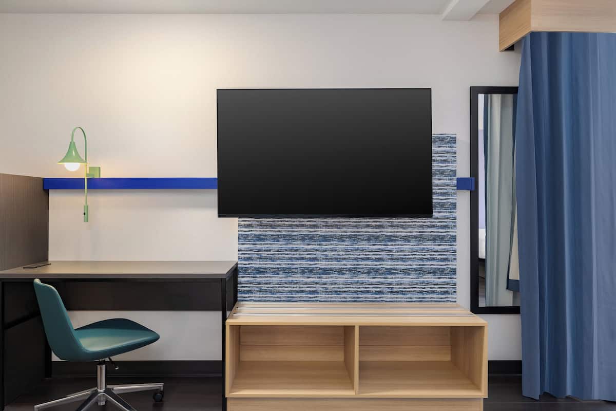 A modern room with a flat-screen TV mounted on a textured wall, a desk with a teal chair, and a wooden shelf below.