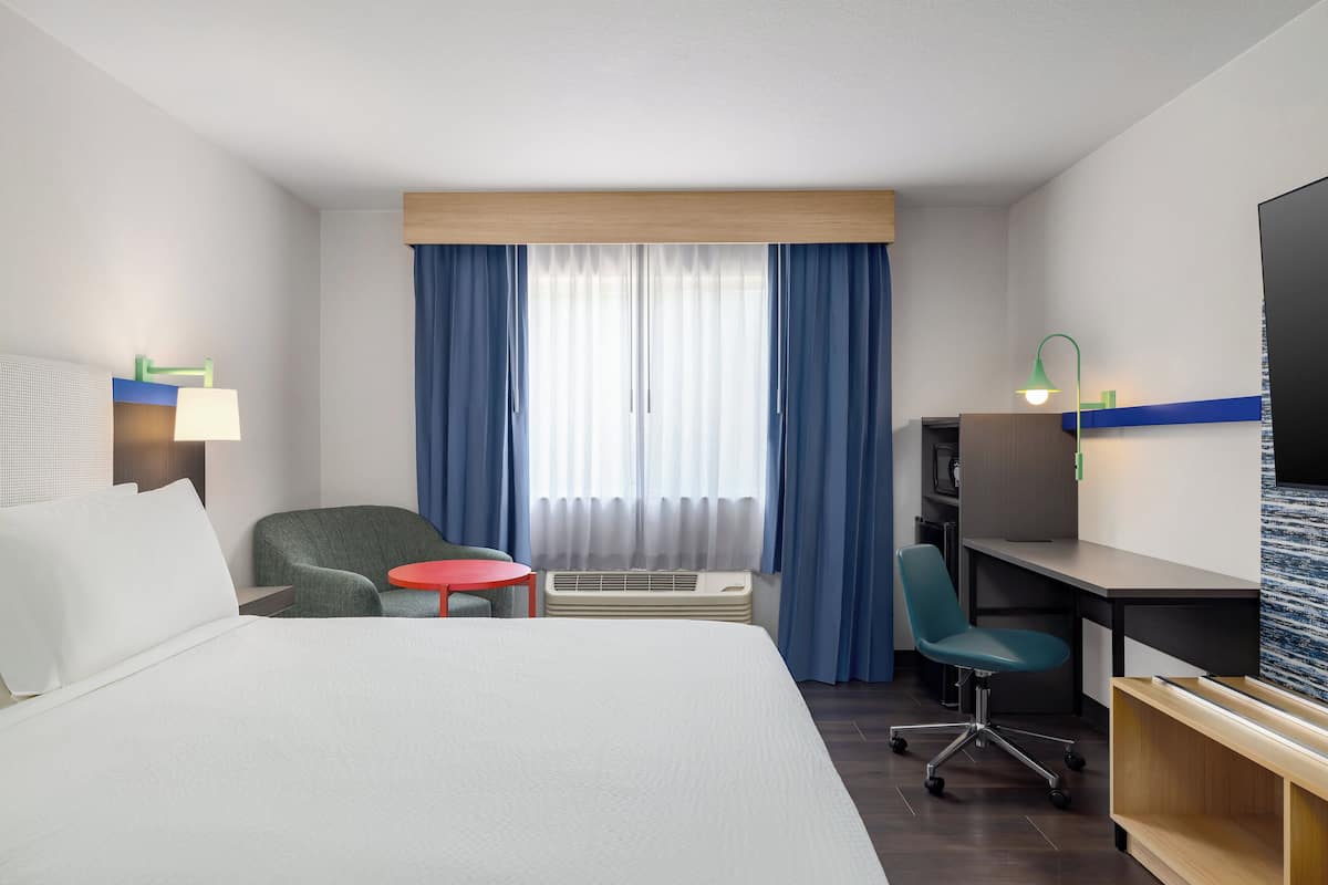 A hotel room with a bed, a desk, a chair, a small table, a TV, and a window with curtains.