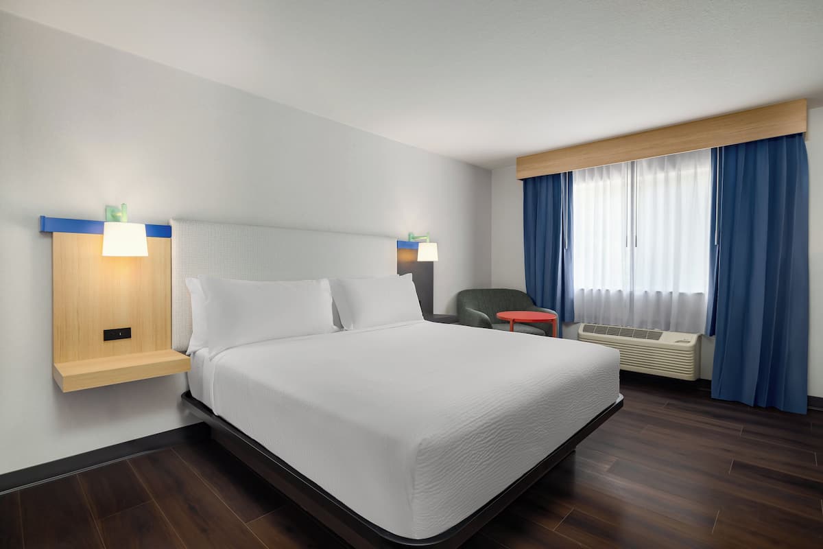 A modern hotel room with a large bed, bedside tables, a chair, a small table, and a window with curtains.