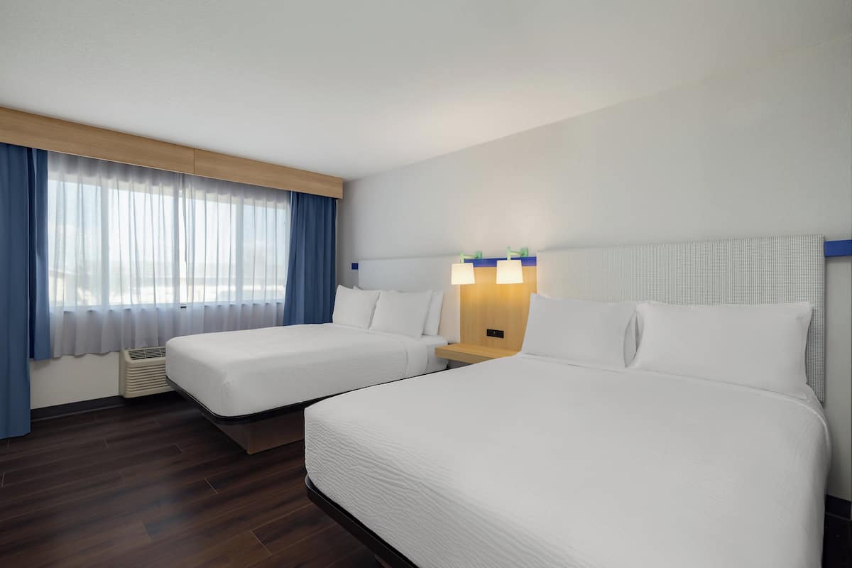A hotel room with two beds, a wooden headboard, a nightstand with lamps, and a window with curtains.