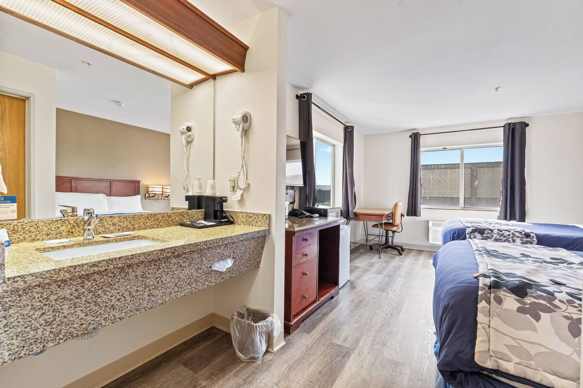 A hotel room with a granite countertop sink, a mirror, a hair dryer, a desk with a chair, a bed with a patterned blanket, and a window with curtains.