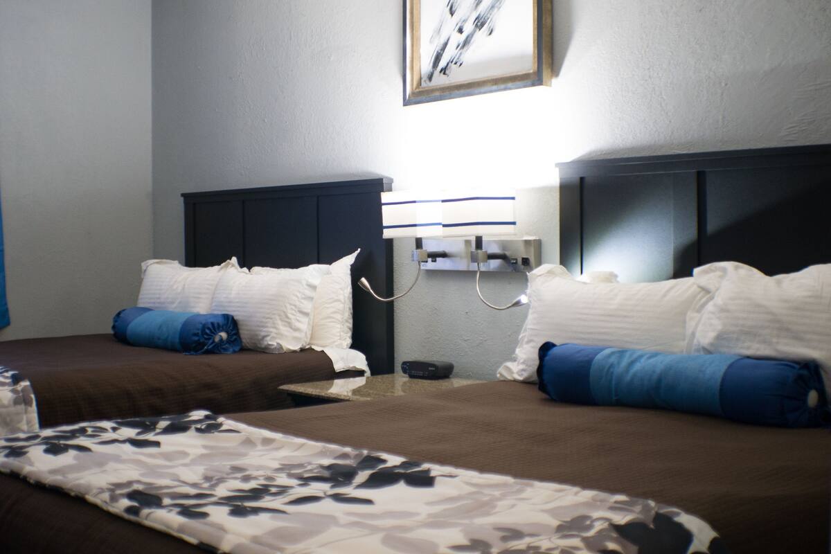 A hotel room with two beds, a bedside table, a wall-mounted lamp, and a framed picture.