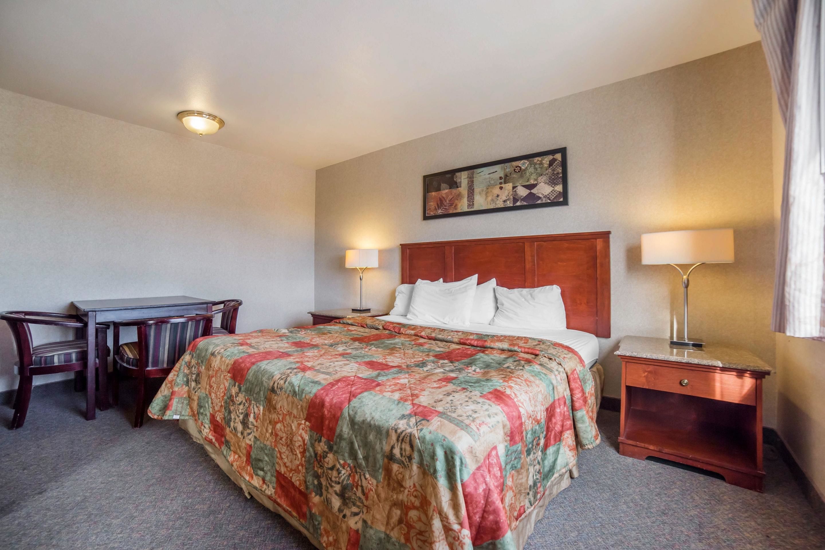 A hotel room with a bed, two bedside tables with lamps, a desk with a chair, and a painting on the wall.