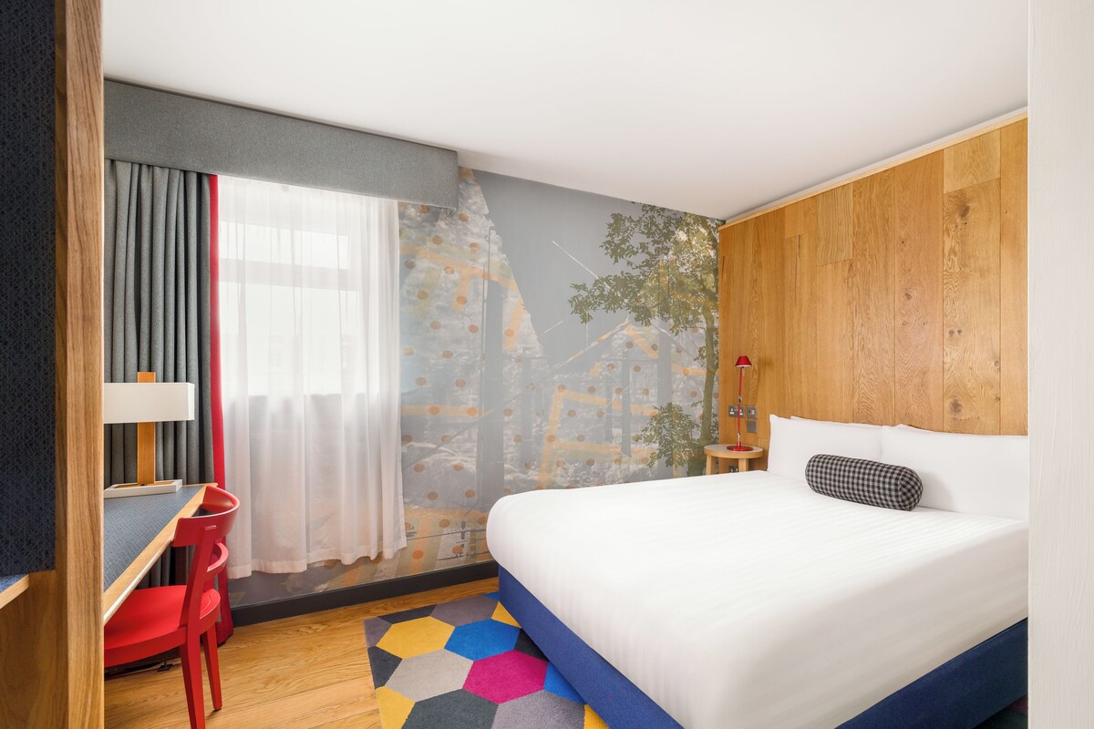 Une chambre d’hôtel avec un lit, un bureau, une chaise, une fenêtre avec des rideaux, un mur orné d’une fresque représentant un arbre et un tapis coloré.
