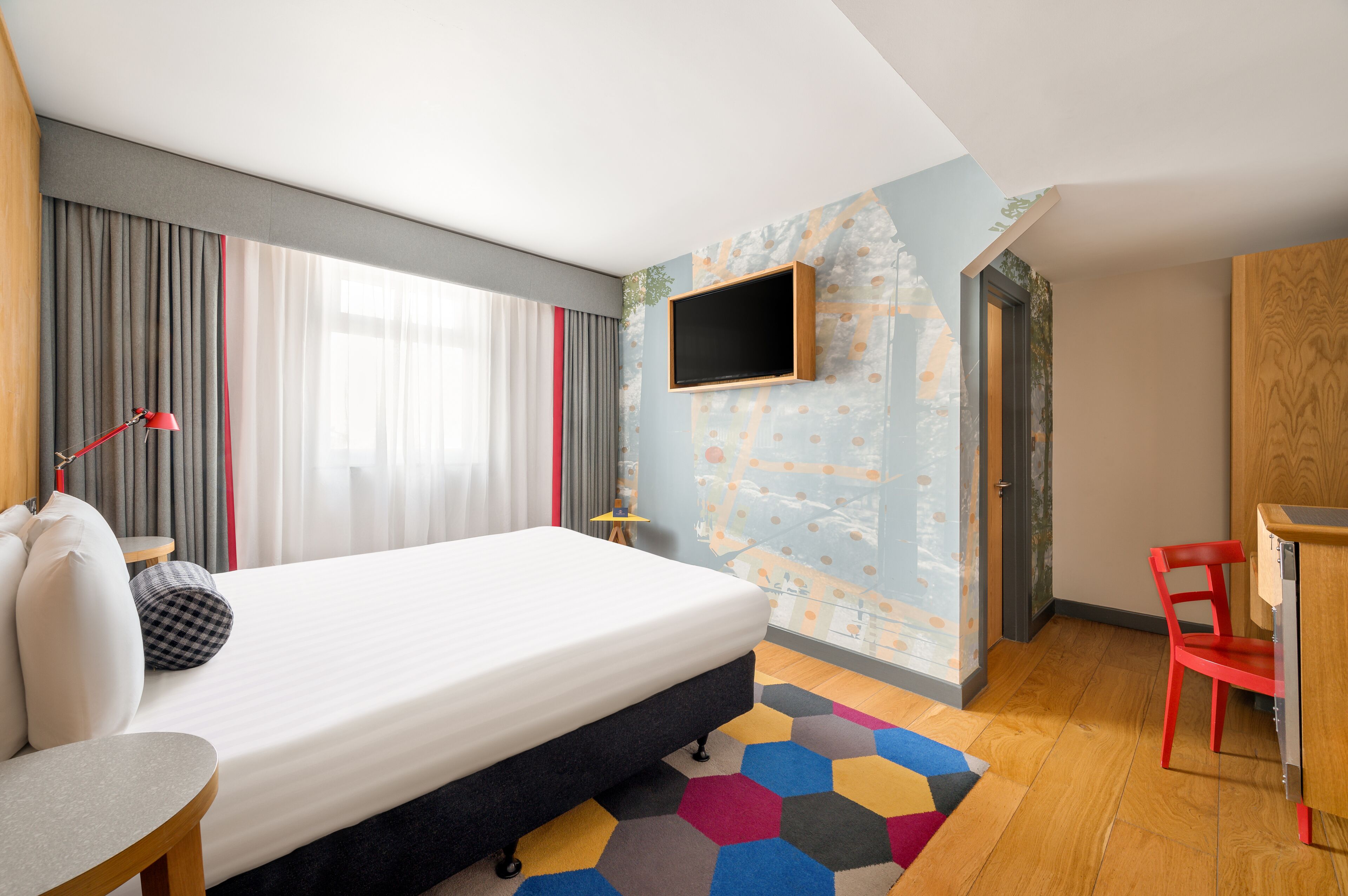 A modern hotel room with a large bed, a TV mounted on the wall, a desk, and a red chair.