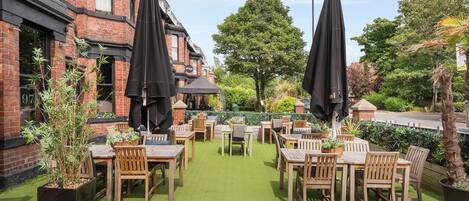 An outdoor patio with wooden tables and chairs, surrounded by brick buildings and greenery.