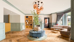 Ein geräumiger Wohnbereich mit einem runden Hocker, einer Couch und einem Couchtisch. In der Mitte befindet sich ein Holzregal und ein Blumentopf.