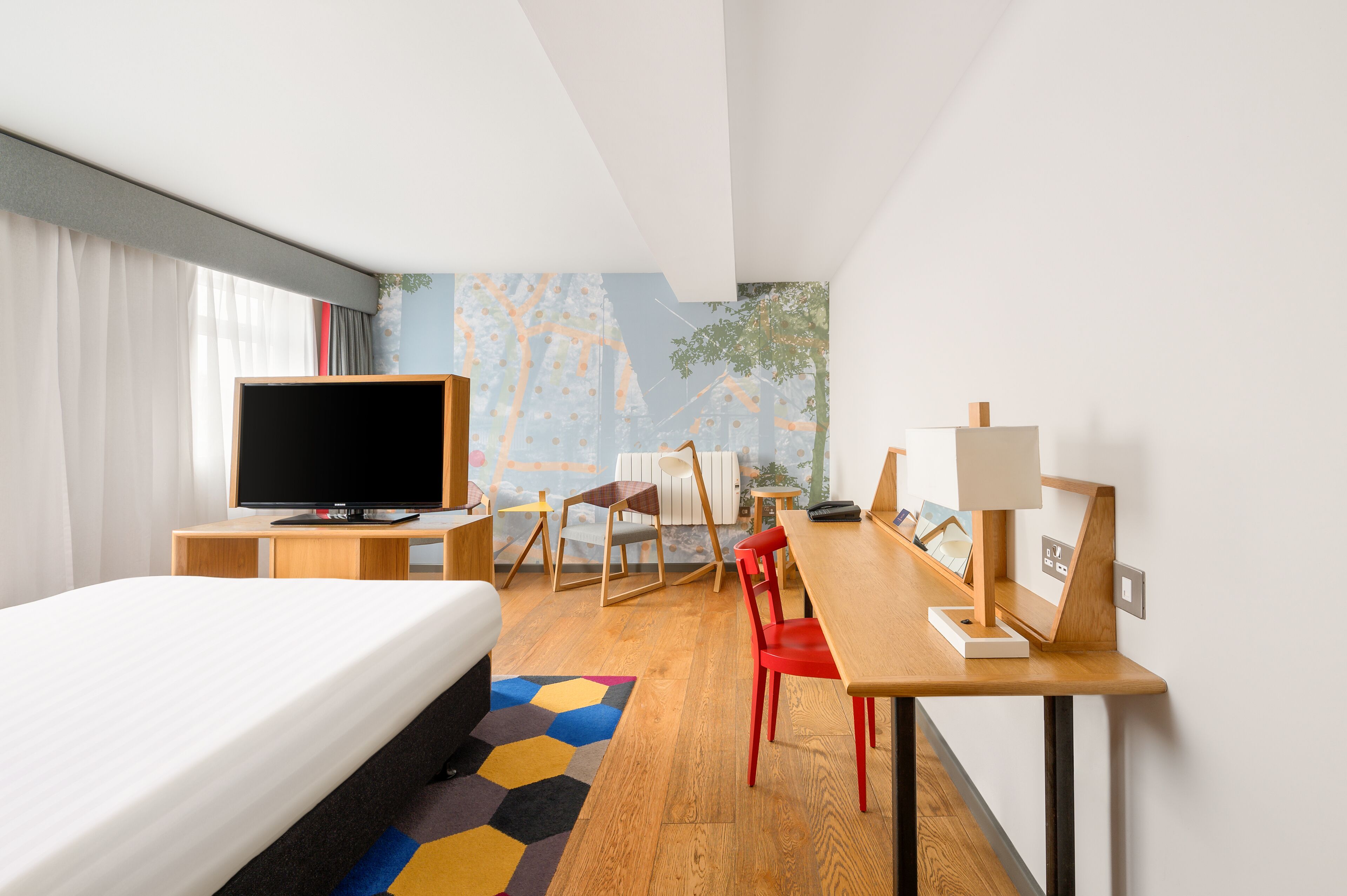 Una habitación de hotel con una cama, un escritorio con lámpara, una silla, un televisor y una alfombra colorida.