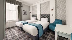 A hotel room with two beds, a desk, a chair, a window with curtains, and patterned wallpaper.