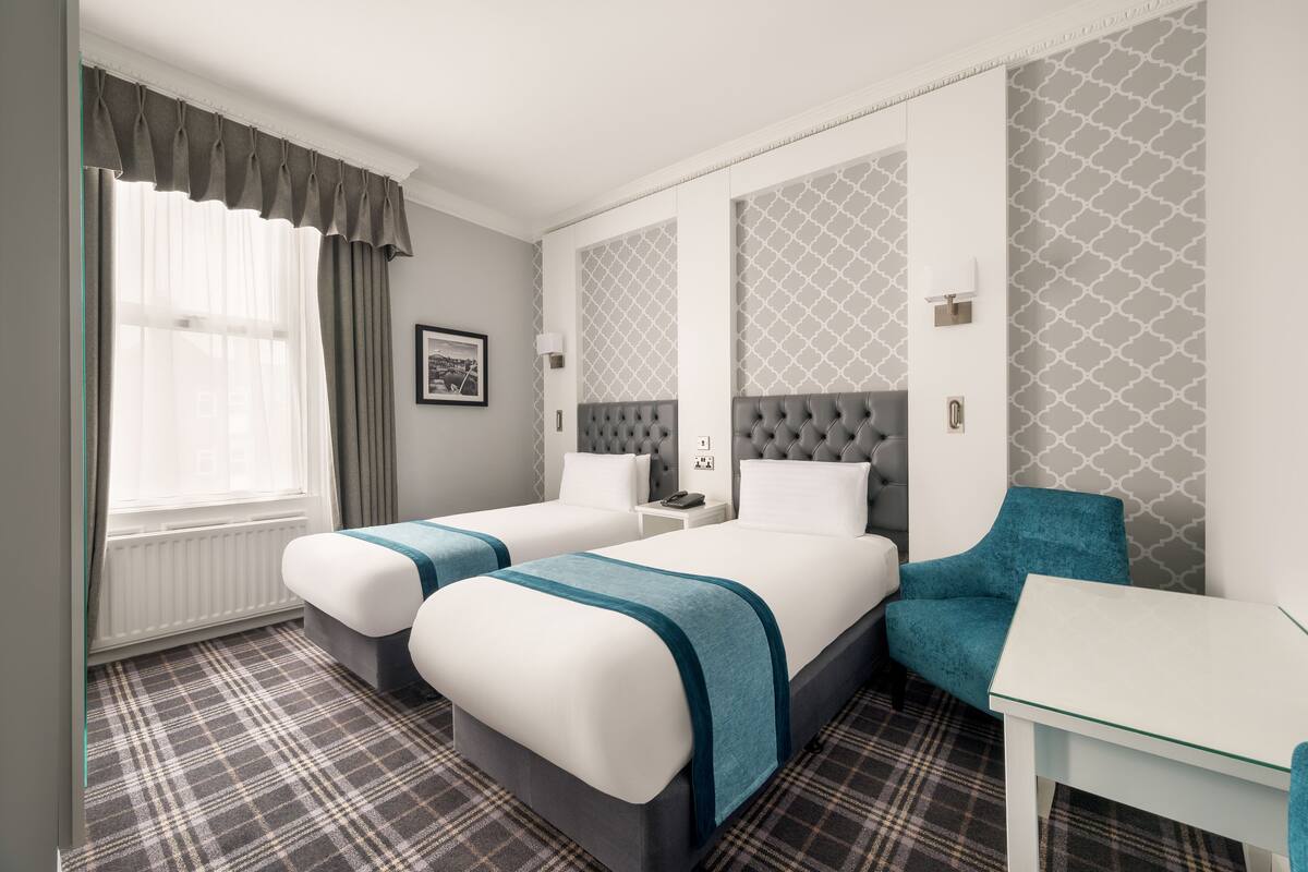 Habitación de hotel con dos camas, una mesita, una silla, un ventanal con cortinas y papel tapiz estampado.