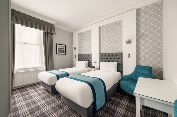Ein Hotelzimmer mit zwei Betten, einem Schreibtisch, einem Stuhl, einem Fenster mit Vorhängen und strukturierter Wandtapete.