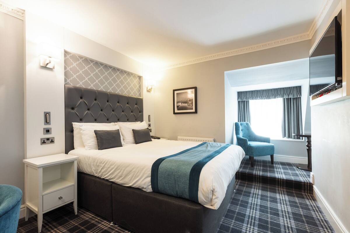 Habitación de hotel con una cama grande, una mesita de noche, una silla, un televisor y una ventana con cortinas.