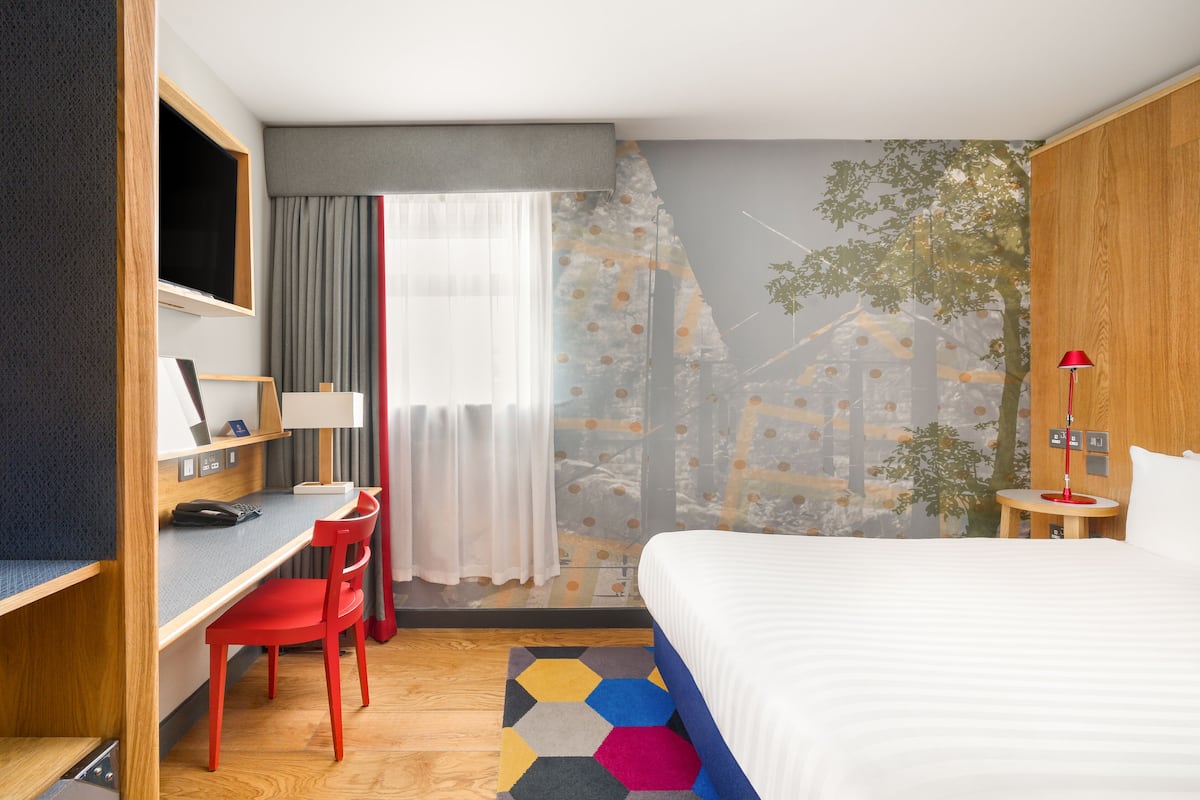 Habitación de hotel con cama, escritorio, silla, televisor y ventana con cortinas.