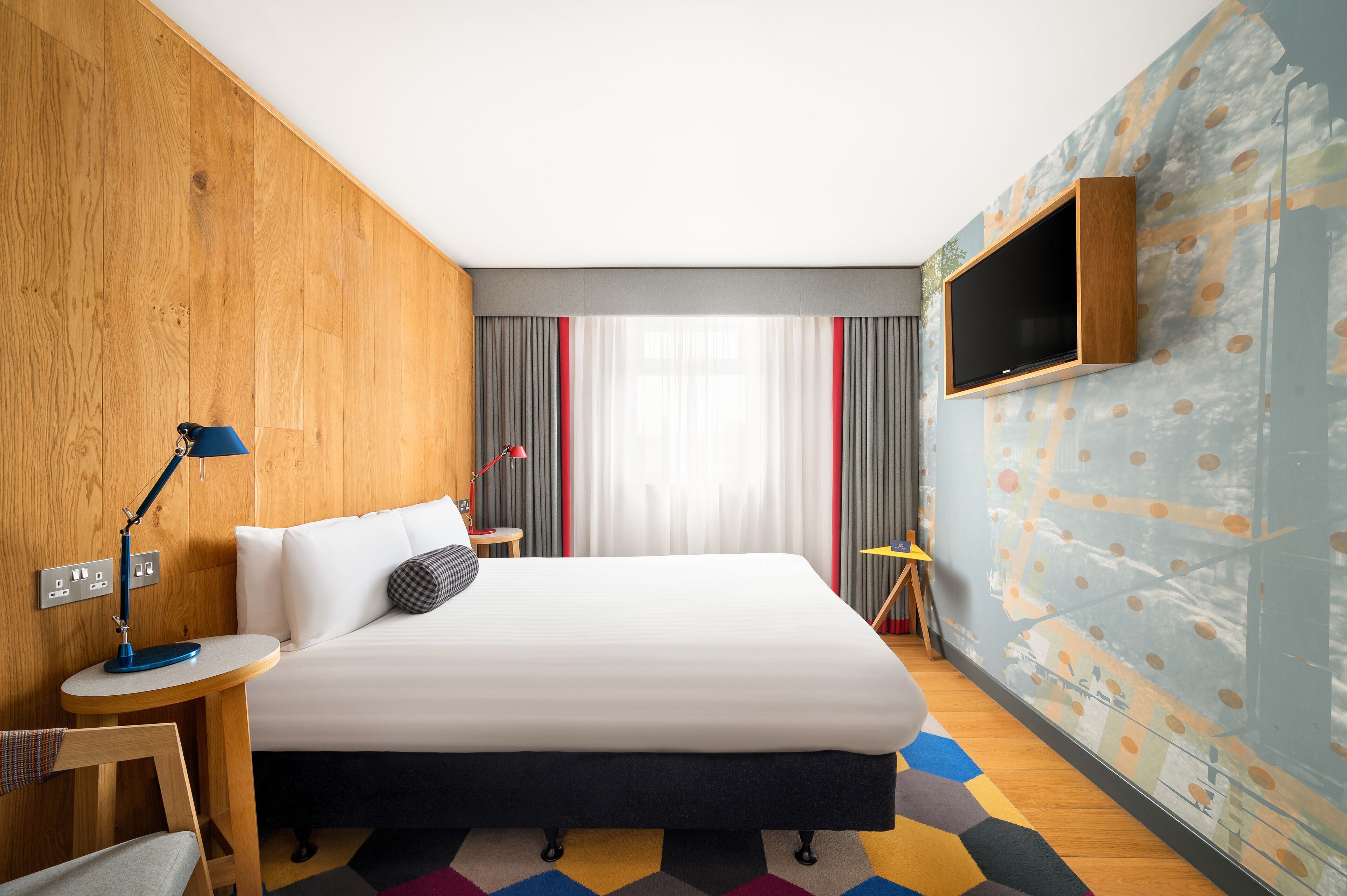 Een moderne hotelkamer met een groot bed, een aan de muur bevestigde televisie, een bureau met een lamp en een kleurrijk vloerkleed.