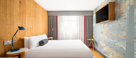 Een moderne hotelkamer met een groot bed, een aan de muur bevestigde televisie, een bureau met een lamp en een kleurrijk vloerkleed.