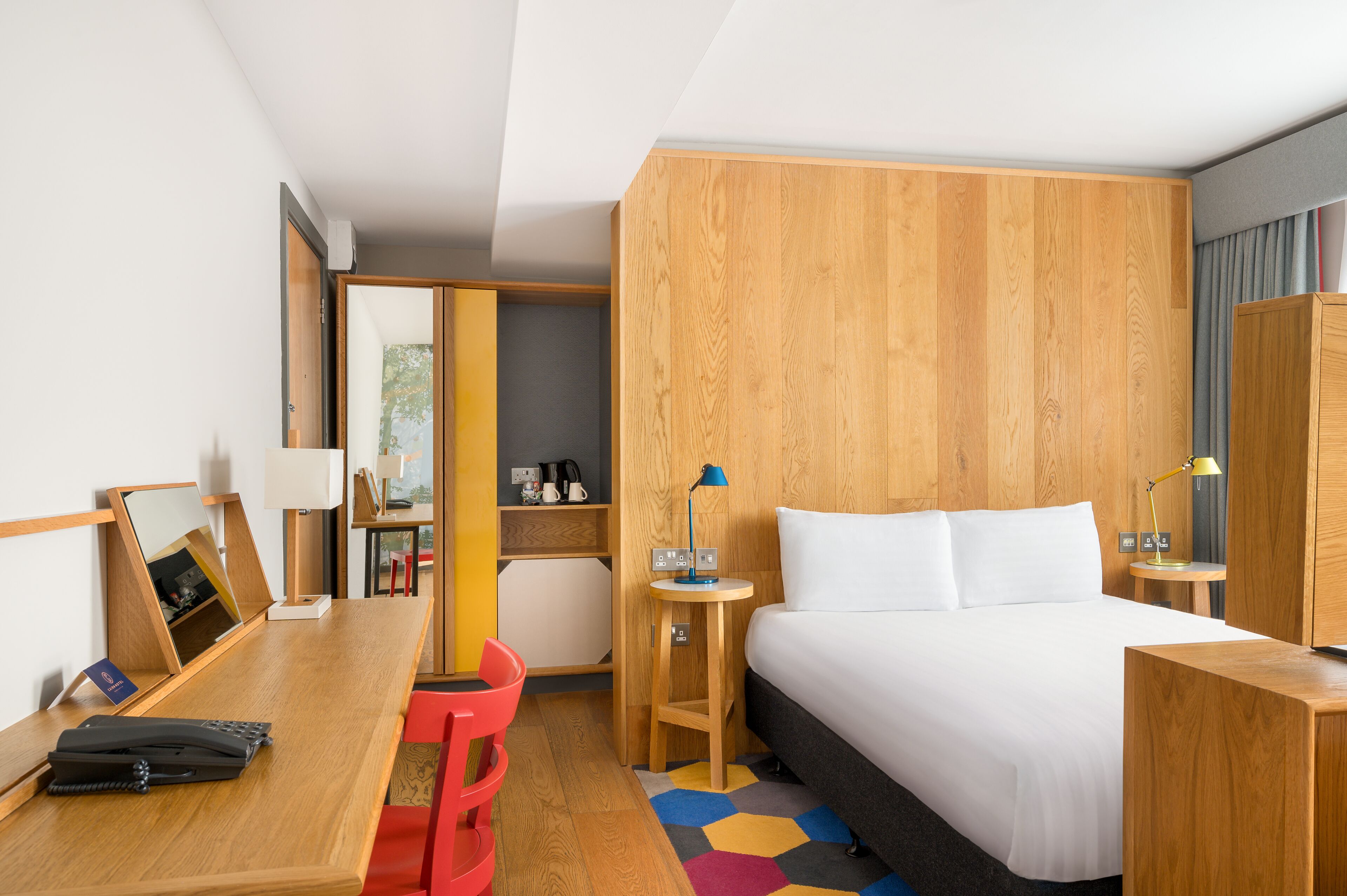 Habitación de hotel con cama de madera, escritorio con silla, mesita de noche con lámpara y armario.