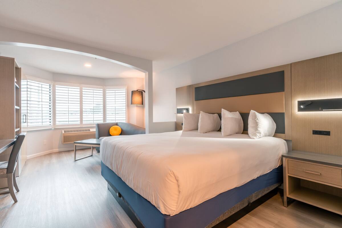 Una habitación de hotel moderna con una cama grande, un escritorio y una silla. Hay una ventana con persianas, una lámpara y un panel de control en la pared.