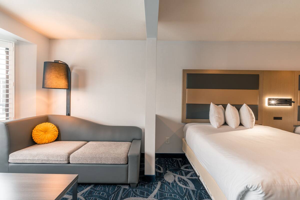 Una habitación de hotel moderna con un sofá gris, una mesita pequeña, una cama con ropa de cama blanca y una alfombra estampada.