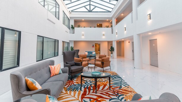 A spacious, well-lit atrium with a high ceiling and skylight, featuring a central seating area with a sofa and armchairs, surrounded by a patterned rug. - Hotel Aluxor SFO (San Bruno)