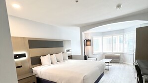 A modern hotel room with a large bed, bedside tables, a desk, and a window with blinds. - Hotel Aluxor SFO (San Bruno)