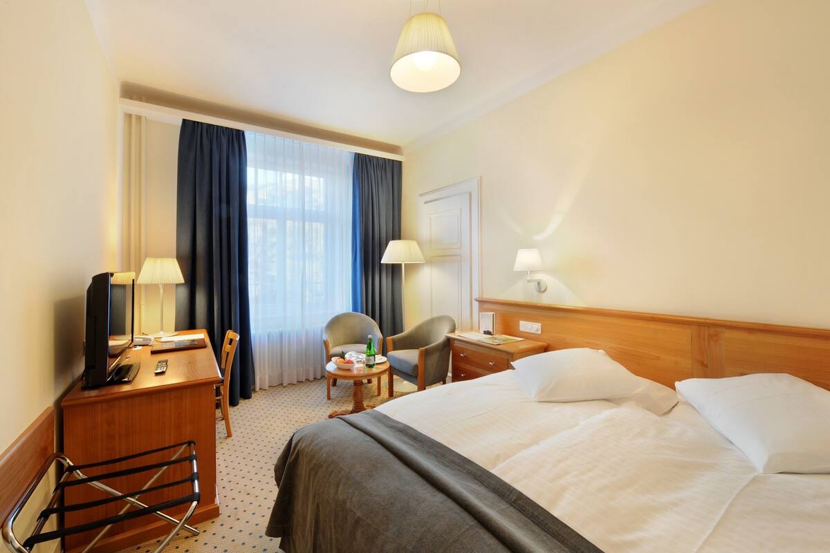 A hotel room with a bed, a desk with a computer, a chair, a small table with a bottle, and a window with curtains.