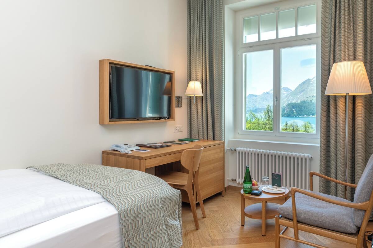 A hotel room with a bed, a desk with a computer, a chair, a TV, and a window with a view of mountains and a lake.