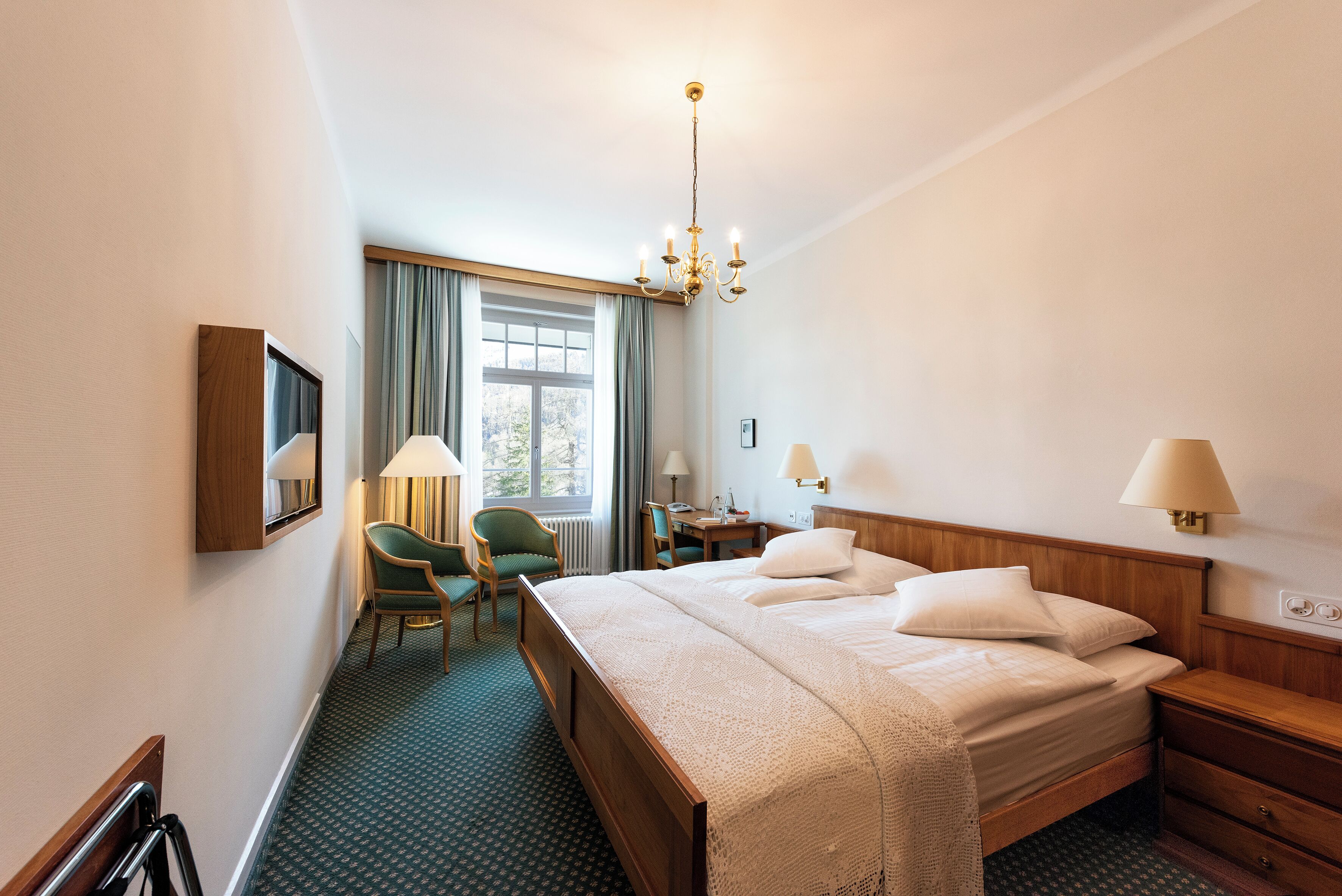 Ein Hotelzimmer mit einem großen Bett, einem Schreibtisch, zwei Stühlen, einer Chandelier und einem fenster mit Vorhängen.