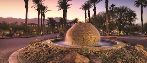 Una fuente de piedra circular con chorros de agua, rodeada por un área ajardinada con rocas, cactus, palmeras y montañas al fondo.