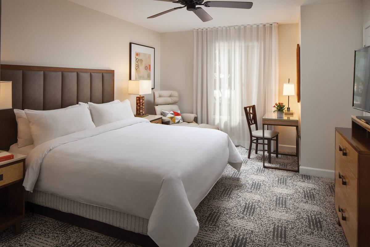 A hotel room with a large bed, a desk, a chair, a ceiling fan, and a television.